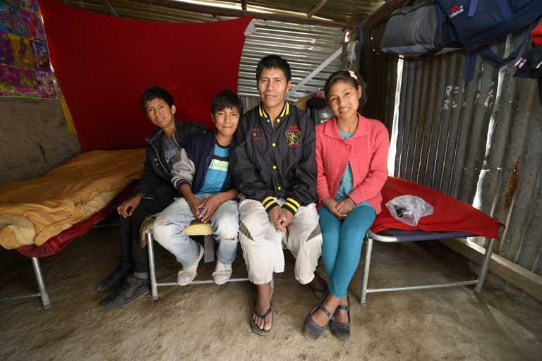 Vater mit Tochter und zwei Söhnen sitzen auf einer Bettkante in einer Blechhütte 