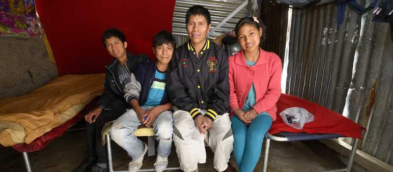 Vater mit Tochter und zwei Söhnen sitzen auf einer Bettkante in einer Blechhütte 